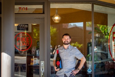 Dane Campbell standing in a doorway holding a large bottle of Pike Road Pinot noir with Pike Road Oregon Crafted painted on the door and address number 701 above the door