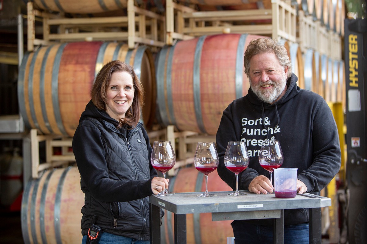 Heather Perkin and Adam Campbell with glasses of Pinot Noir samples in front of barrels