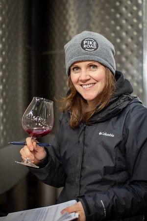 Pike Road Head Winemaker Heather Perkin holding a glass of wine and a clipboard in front of steel fermentation tanks