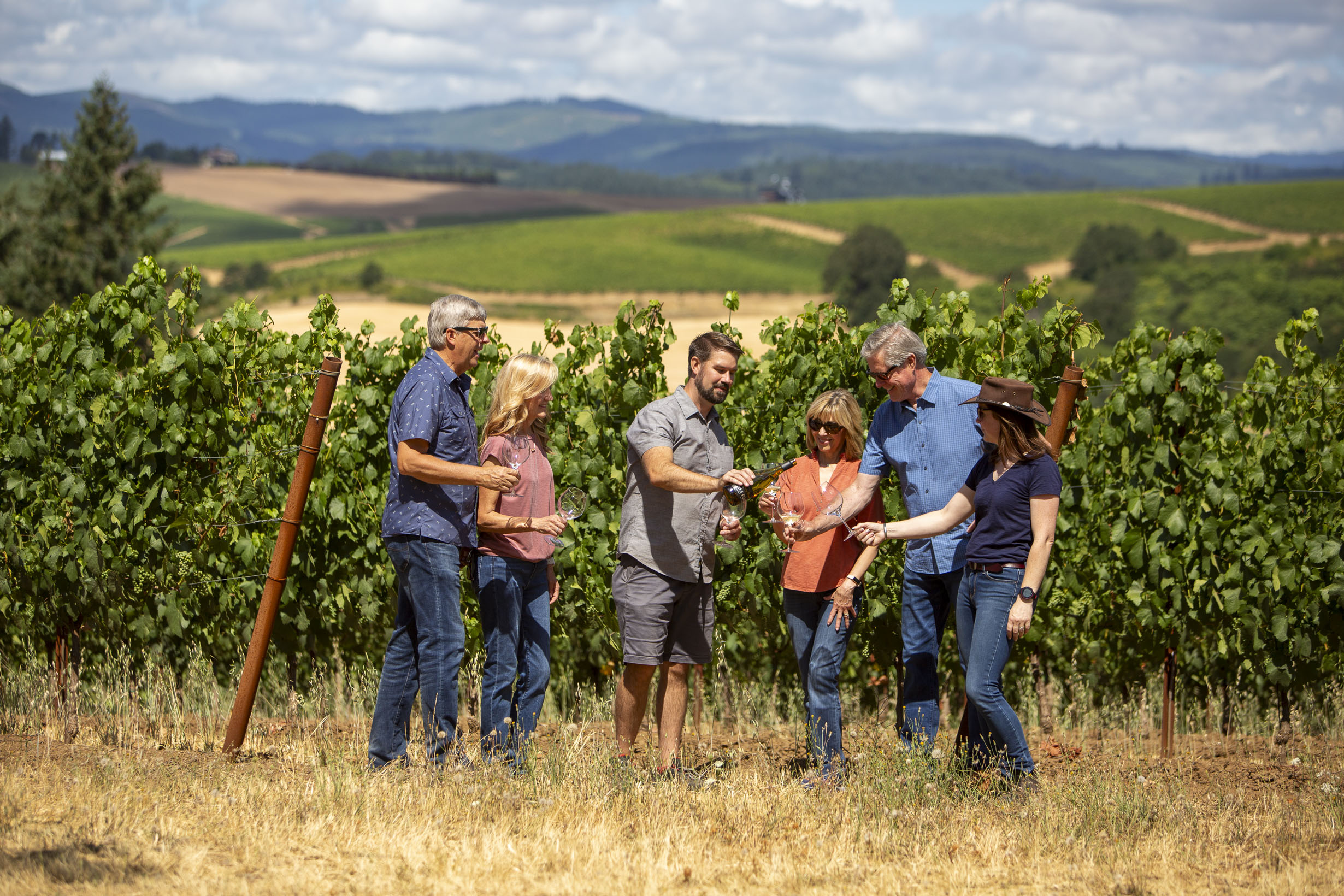 a group of people standing in a vineyard and pouring red wine